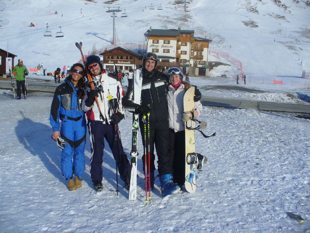 Vier Personen posieren mit Skaterpads und Werkzeugen vor einer Schnee- und Eispiste mit Gebäuden und anderen Menschen im Hintergrund.