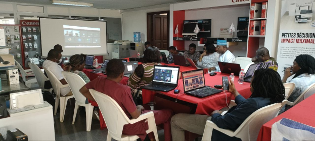 Gruppe von Menschen an einem Tisch mit Laptops, Flaschen und anderen Gegenständen während eines digitalen Marketing-Workshops, mit einem Banner, einem Display-Bildschirm, einer Tür und Deckenleuchten im Hintergrund.