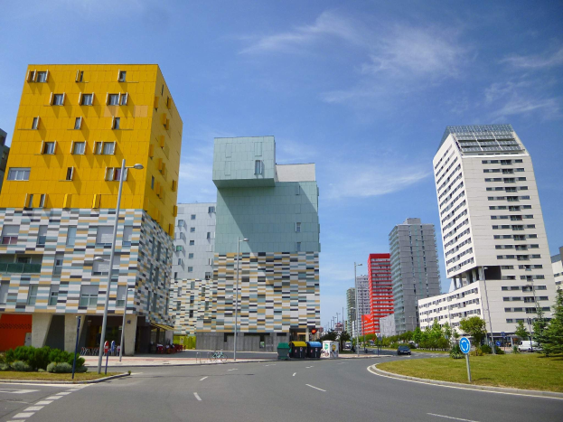 Stadtstraße mit hohen Gebäuden der Universität Rotterdam, Straßenlaternen, Lichtern, Fahrzeugen, Infotafeln, Grünflächen und bewölktem Himmel.