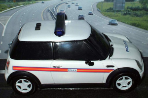 Ein weißer Polizeiwagen ist im Vordergrund mit mehreren Autos, die sich auf der Straße dahinter bewegen, umgeben von Gras und Bäumen auf beiden Seiten, in einer ländlichen Umgebung.