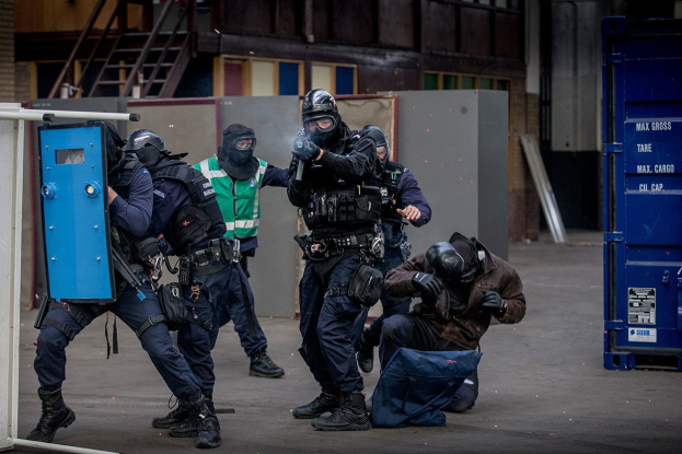 Eine Gruppe von Polizisten in Schutzausrüstung steht vor einem Gebäude in einer Reihe, wobei eine Person im Vordergrund liegt und Metallgegenstände, Treppen und eine Wand im Hintergrund zu sehen sind.