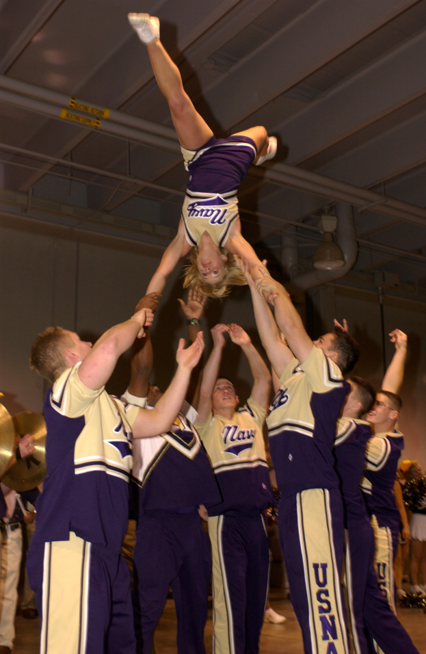 Eine Gruppe von Cheerleadern führt einen Stunt in einer Turnhalle durch, mit einer Frau in einem Handstand in der Mitte, anderen Umstehenden, einigen mit Musikinstrumenten auf der linken Seite und einer Wand mit Rohren und Deckenleuchten im Hintergrund.