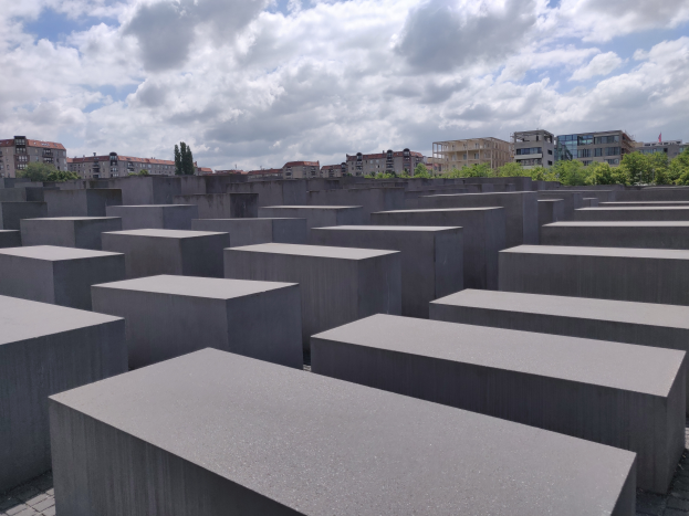 Ein großes Feld aus Betonblöcken in verschiedenen Höhen, die in einem Gittermuster angeordnet sind, mit Bäumen und Gebäuden im Hintergrund und einem bewölkten Himmel darüber.