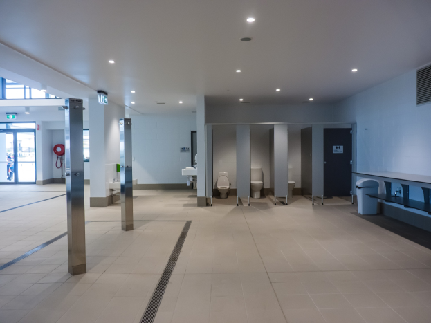 Große Öffentliche Toilette mit mehreren Toiletten, tragenden Säulen, einem Mülleimer, einer Tür, einer Tafel, einem Feuerlöscher und deckenmontierten Leuchten.
