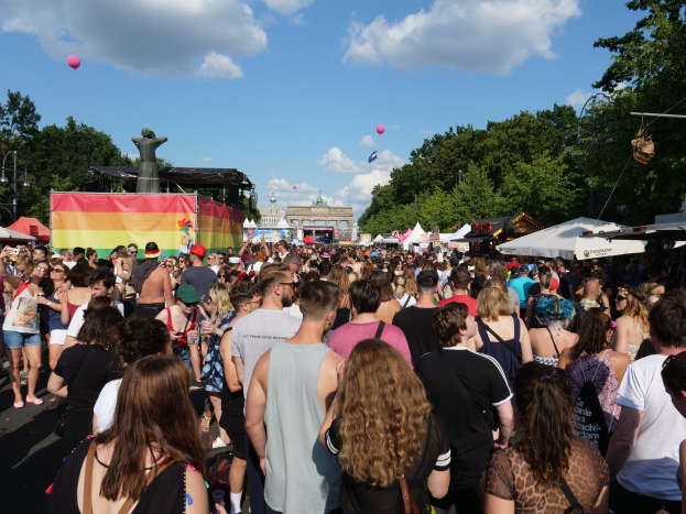 Eine große Menschenmenge, die eine Straße mit Zelten, Bäumen, Pfählen und Lichtern entlanggeht, mit Gebäuden und einem bewölkten Himmel mit Ballons im Hintergrund beim Berlin Pride Festival.