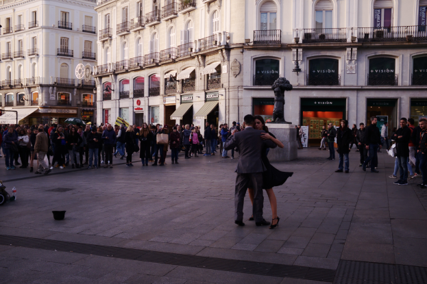 Ein Mann und eine Frau tanzen auf einem belebten Stadtplatz, umgeben von Zuschauern, mit einem Kinderwagen, einem Eimer und Stangen links und Gebäuden, Schildern, einer Statue und einem bewölkten Himmel im Hintergrund.