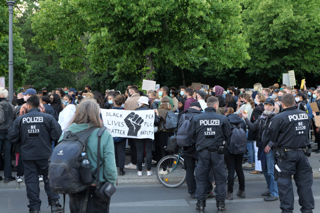 Eine große Gruppe von Menschen steht auf der Straße, einige halten Schilder, mit einem Fahrrad im Vordergrund und Bäumen und einem Pfahl im Hintergrund, bei einer Black-Lives-Matter-Demonstration in Berlin.