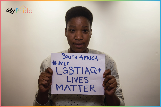 Eine Frau in einem weißen Hemd steht entschlossen vor einer Wand und hält ein Schild mit der Aufschrift "LGBTQ+ Lives Matter" in großen schwarzen Buchstaben hoch.