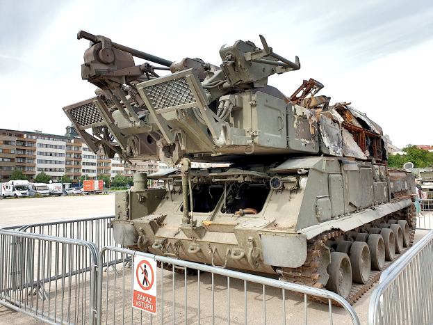 Ein gepanzerter Transporter vor einem Metallzaun mit einem Schild daran geparkt, mit Fahrzeugen, Bäumen, Gebäuden und einem klaren blauen Himmel im Hintergrund.