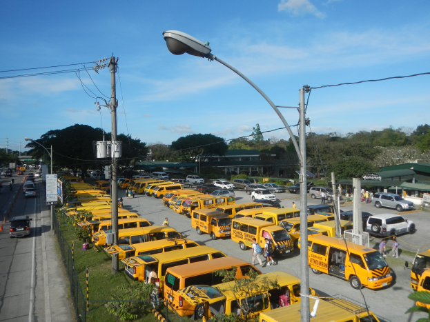 Eine große Anzahl gelber Schulbusse, die am Straßenrand geparkt sind, mit Menschen, die auf dem Gehweg gehen, Strommasten mit Drähten, Laternen, Bäume, Gebäude und ein bewölkter Himmel im Hintergrund.