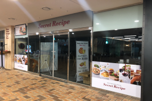 Der Eingang zu einem Restaurant namens 'Geheimes Rezept' mit Glastüren, einem Stand mit verschiedenen Gegenständen und einer Speisekarte auf der linken Seite und Deckenleuchten über den Türen.