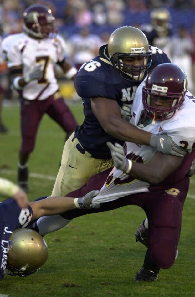 Zwei Fußballer mit Helmen und Uniformen stoßen auf dem Feld zusammen, während andere im Hintergrund zuschauen.
