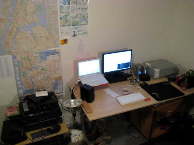 Ein Raum mit einem Tisch, auf dem ein Laptop, ein Lautsprecher, ein Monitor, eine Tastatur, ein Ball, eine Maus, ein Scanner und Papiere stehen, und einem weiteren Tisch mit Elektronikgeräten im Hintergrund, mit Charts und Postern an der Wand dahinter.
