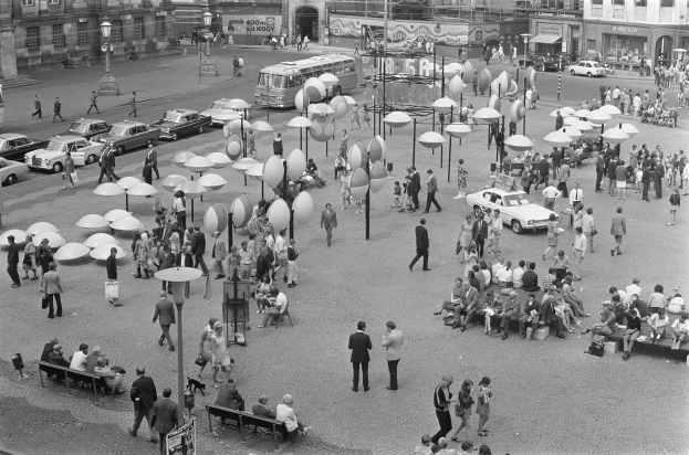 Ein Schwarz-Weiß-Foto eines belebten Stadtplatzes mit Menschen, die gehen, auf Bänken sitzen, Laternen und Fahrzeugen sowie Gebäuden mit Fenstern und Schildern im Hintergrund.