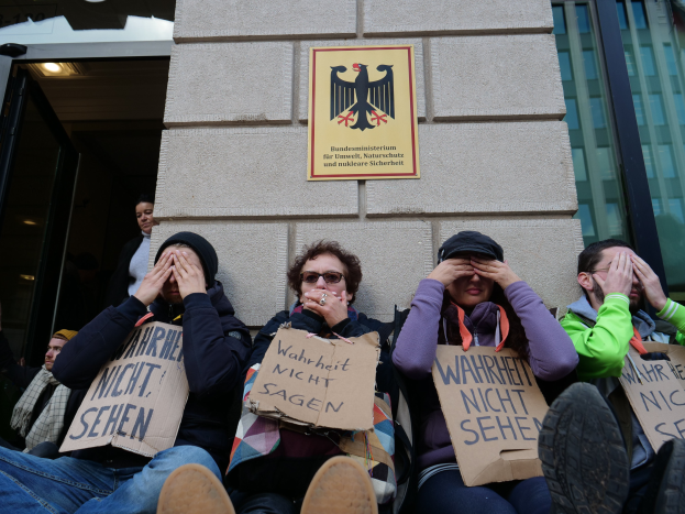 Eine Gruppe von Menschen sitzt vor einem Gebäude und hält Schilder, einige bedecken ihre Gesichter, mit einem Logo und einer Tafel an der Wand und einer Glastür auf der rechten Seite.