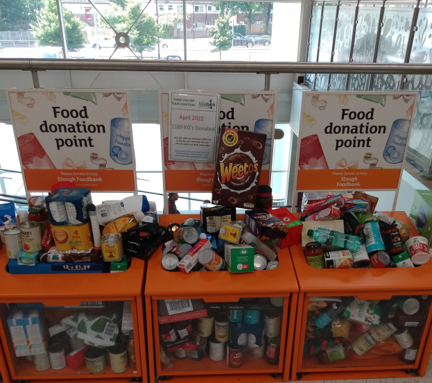 Orange Schübe mit Lebensmitteln wie Flaschen, Dosen und Kisten an einer Spendenstelle, mit Informationsschildern und einer Aussicht auf Fahrzeuge, Bäume, Gebäude und Himmel durch Glasfenster.