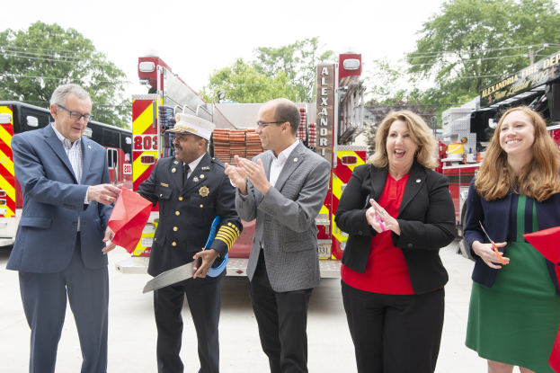 Eine Gruppe von Menschen steht vor einem Feuerwehrauto, klatscht und lächelt, mit zwei Personen, die eine Schere und ein rotes Tuch halten, während einer Einweihungszeremonie der Feuerwehr Alexandria, mit Bäumen, Drähten und einem klaren blauen Himmel im Hintergrund.