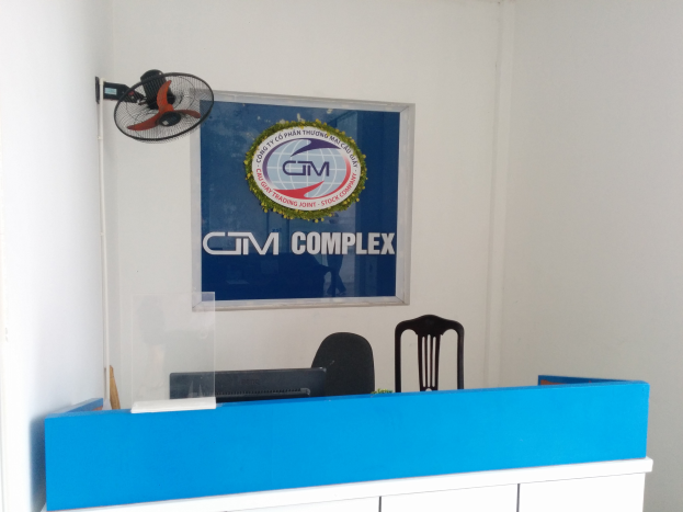 Ein blauer und weißer Empfangstisch mit einem Computer, umgeben von Stühlen und einem Tischventilator, mit einer Tafel an der Wand dahinter, die Text und ein Logo anzeigt, was darauf hinweist, dass es das Büro von GM Trading Joint in Ho Chi Minh Stadt ist.