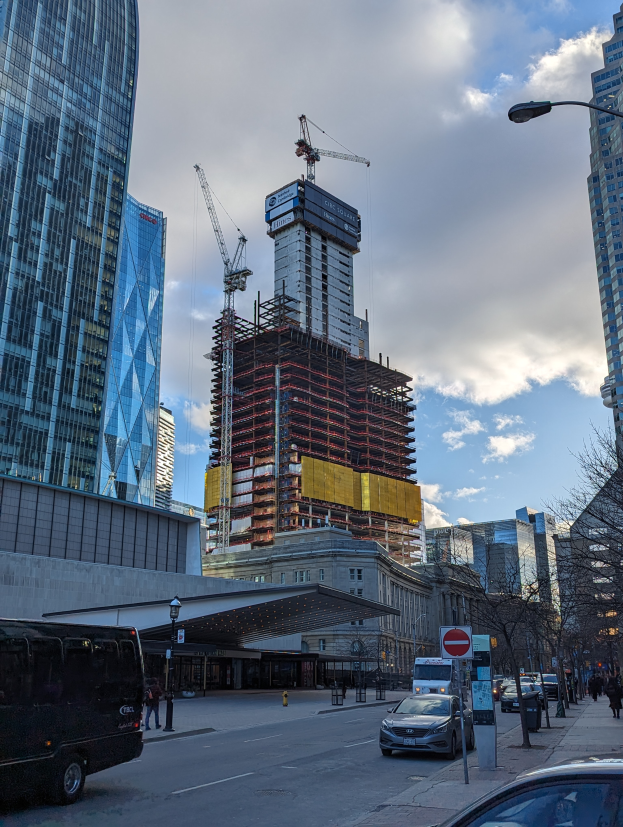 Eine belebte Straße mit Fahrzeugen, Passanten, Schildern, Laternenmasten und Bäumen, mit dem One World Trade Center im Hintergrund unter einem bewölktem Himmel.