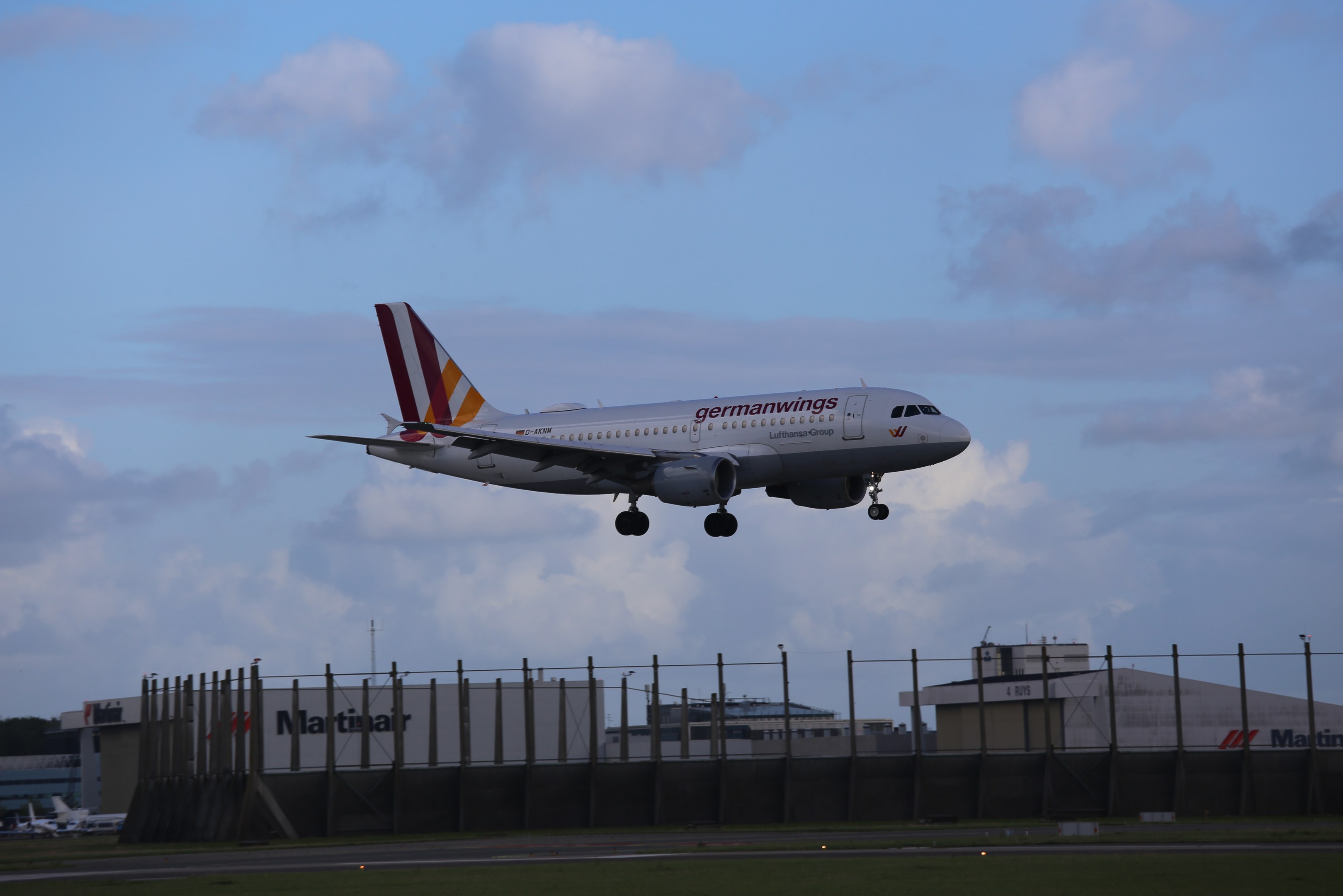 Germanwings Airbus A320-200 beim Start vom Frankfurt Airport, mit Gras darunter, einem Zaun im Vordergrund, Gebäuden und Bäumen im Hintergrund und einer bewölkten Himmel über.