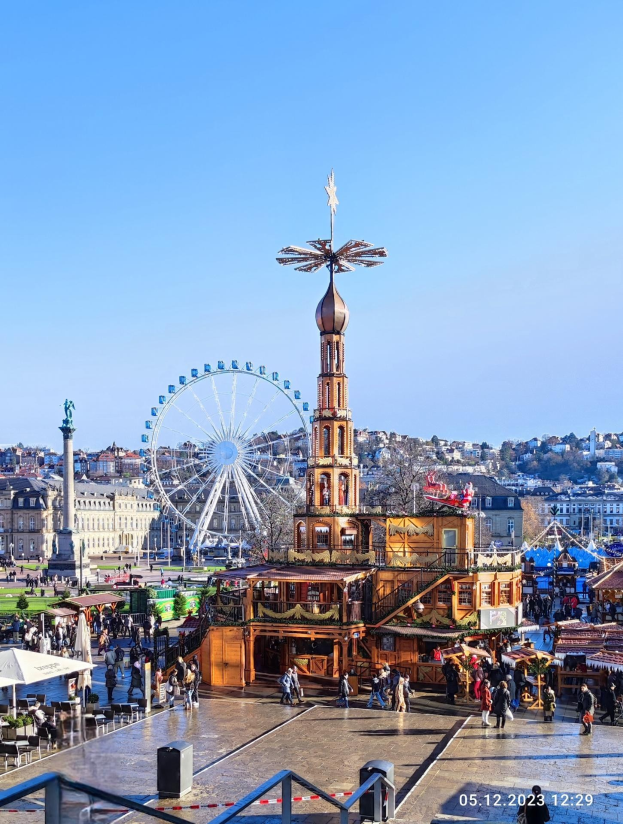 Ein belebter Stadtplatz mit einem zentralen Riesenrad, umgeben von Gebäuden, Bäumen und Menschen unter einem klaren blauen Himmel, mit Zelten und Geländern im Bild.