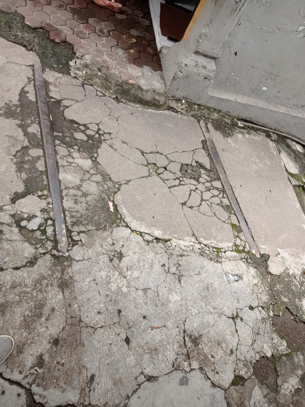 Eine Person steht auf einem gebrochenen Gehweg neben einem Gebäude, mit einem Metallobjekt auf dem Boden, einer Wand und einer Tür sichtbar.