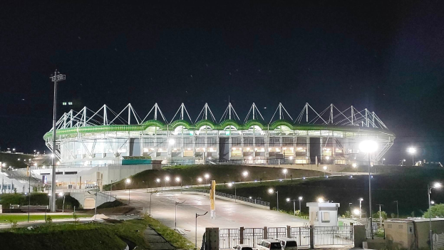 Ein großes Stadion bei Nacht beleuchtet, umgeben von Straßenmasten, Straßenlaternen, Strommasten, Stromkabeln, parkenden Autos, Fahrzeugen auf der Straße, Grills, Bäumen und einem sternenklaren Himmel.
