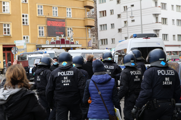 Polizeibeamte in Uniform vor einer Menge von Helmträgern während einer Demonstration in Berlin, mit Fahrzeugen, Gebäuden und einem Kameramann im Bild.