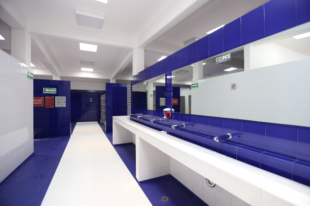 Öffentliche Toilette mit blauen Kacheln an den Wänden, weißen Arbeitsplatten, Waschbecken mit Wasserhähnen, einer Plattform mit einer Flasche und anderen Gegenständen, Texttafeln an den Wänden, Deckenleuchten und Säulen.