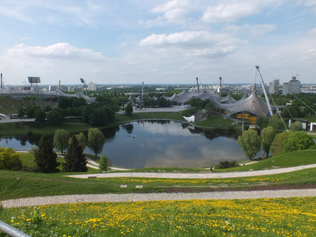 Ansicht des Olympiastadions von einem Hügel aus, mit einem Teich im Vordergrund, umgeben von Grün und gelben Blumen, und Gebäuden im Hintergrund unter einem klaren blauen Himmel.