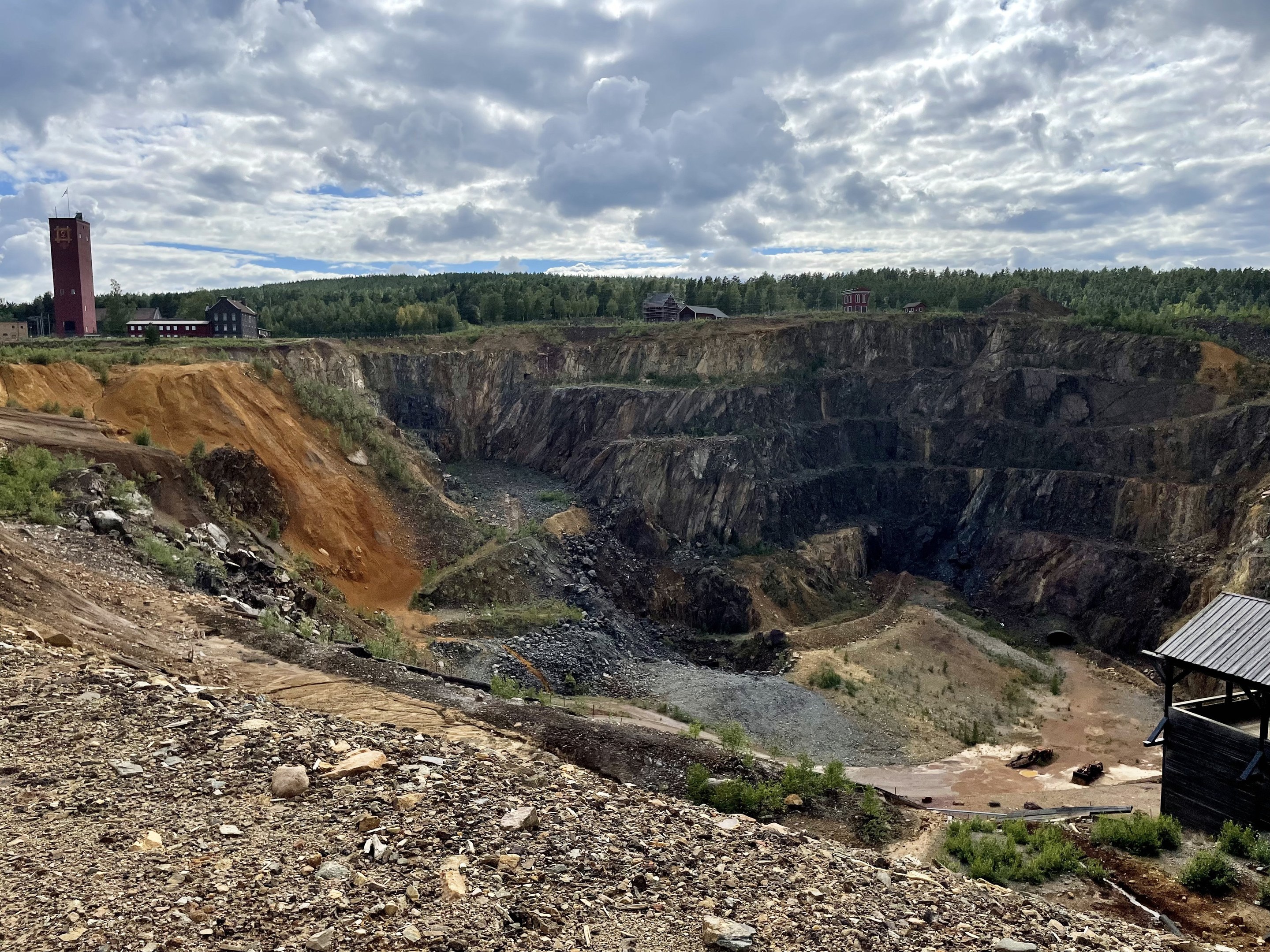 Großer Tagebau in einem Schmutzfeld umgeben von Bäumen, Gebäuden und einem Schuppen unter einem bewölkten Himmel, mit Steinen und Pflanzen auf dem Boden.
