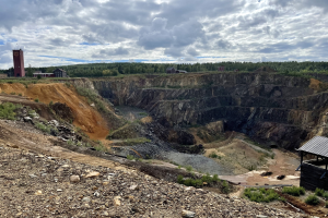 Großer Tagebau in einem Schmutzfeld umgeben von Bäumen, Gebäuden und einem Schuppen unter einem bewölkten Himmel, mit Steinen und Pflanzen auf dem Boden.