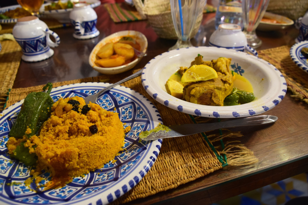 Ein Tisch mit Tellern, Gläsern, Schüsseln, Krügen, Besteck und Matten, mit Menschen im Hintergrund bei einem traditionellen marokkanischen Essen.