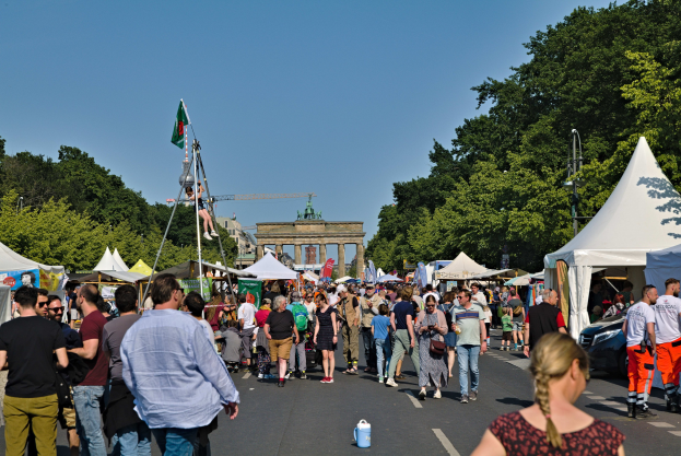Menschenmenge auf einer Straße mit Zelten, Fahrzeugen und Bäumen, im Hintergrund ein Tor, eine klare blauer Himmel und Fahnenmäste
