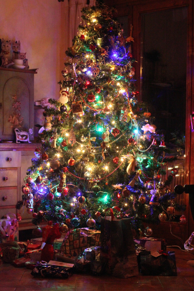 Ein Weihnachtsbaum mit Lichtern und Schmuck, Geschenke auf dem Boden, eine Vitrine mit Gegenständen links und ein Fenster an der rechten Wand.