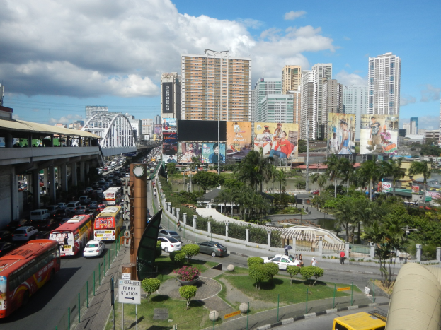 Eine belebte Stadtstraße mit Bussen, Autos und anderen Fahrzeugen, gesäumt von Bäumen, Gebäuden und einer Brücke unter einem bewölkten Himmel.