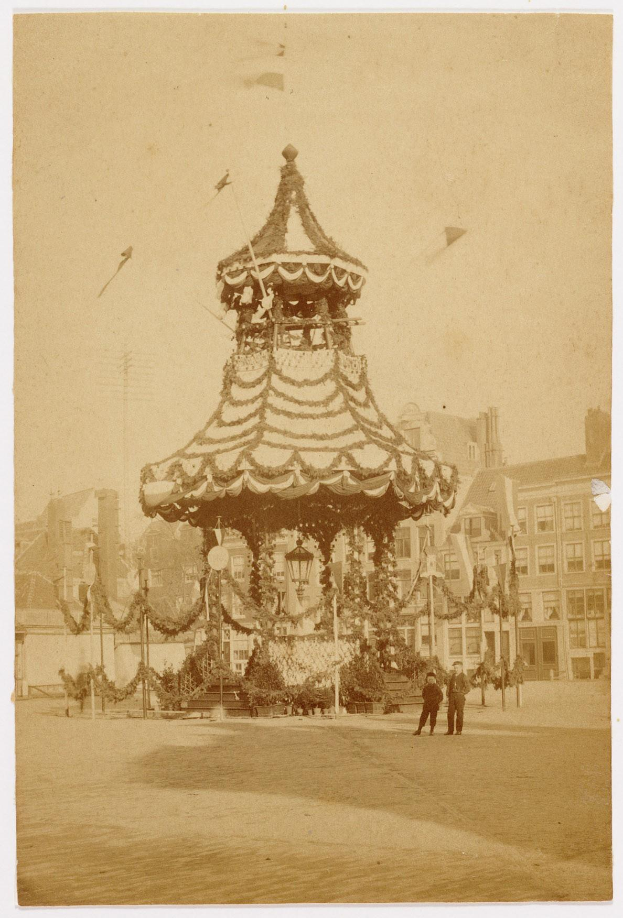 Ein altes Schwarz-Weiß-Foto eines Stadtkarussells, umgeben von Menschen, Pfählen, Lichtern und Gebäuden, geschmückt mit festlichen Weihnachtsdekorationen, mit dem Himmel im Hintergrund.