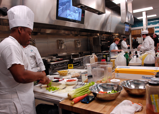 Eine Gruppe von Köchen in einer Küche beim Zubereiten von Speisen auf einem Schneidbrett, wobei ein Koch im Vordergrund in einer weißen Schürze und Mütze steht und ein Messer hält und von Utensilien, Schüsseln, Gemüse, einem Mobiltelefon, Flaschen und anderen Gegenständen umgeben ist, mit einem Fernseher und Deckenlampen im Hintergrund.