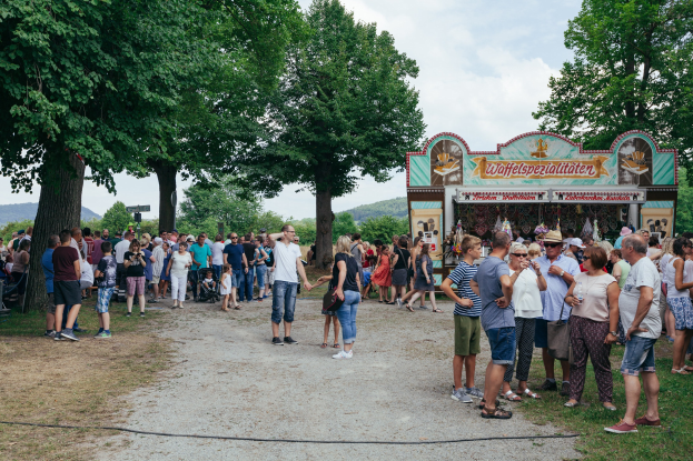 Eine Menschenmenge steht vor einem Essensstand auf dem Oktoberfest in München, Deutschland, einige tragen Mützen und Okulare, andere halten Gegenstände, vor einem grasbewachsenen Bereich mit Bäumen, Hügeln und einem bewölkten Himmel.