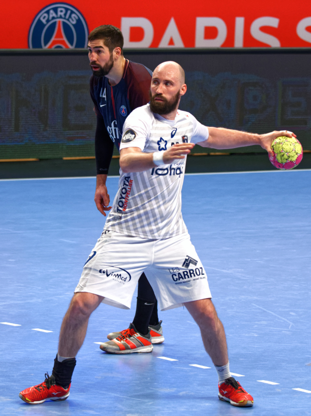Zwei Männer spielen Handball auf einem Platz, einer hält einen Ball, mit einer Tafel im Hintergrund, auf der "Paris Saint-Germain vs Paris Saint Germain" steht.