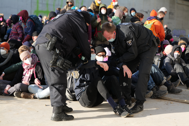 Eine Gruppe von Menschen, einige mit Mützen und Masken, sitzt auf dem Boden mit zwei Polizeibeamten in der Nähe in einer Stadtumgebung, mit einer Wand mit Schrift im Hintergrund.
