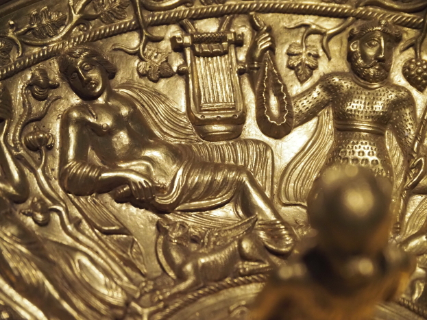 Nahaufnahme einer Metallskulptur, die eine Frau in einem langen, wallenden Kleid mit einem Stab und einem Schild und einen Mann in Rüstung mit einem Objekt darstellt, beide von einem Heiligenschein umgeben.