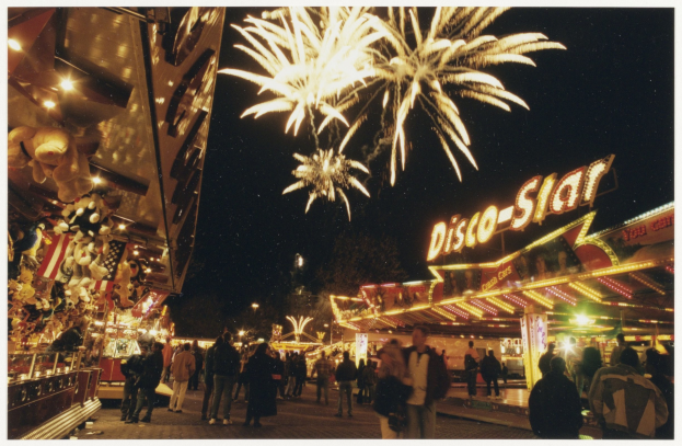 Eine Menschenmenge steht nachts vor einer Karussellfahrt, beleuchtet von Lichtern und umgeben von Gebäuden mit Schildern, mit Bäumen und Feuerwerk im Hintergrund.