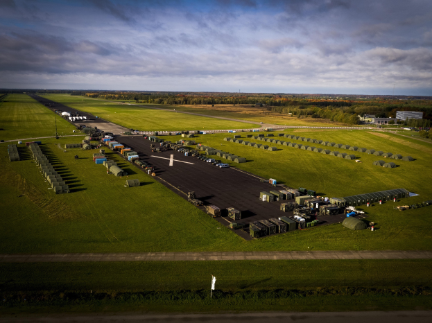 Luftaufnahme einer Militärbasis in den Niederlanden mit Straßen, Fahrzeugen, Gras, Bäumen, Gebäuden und einem bewölkten Himmel.