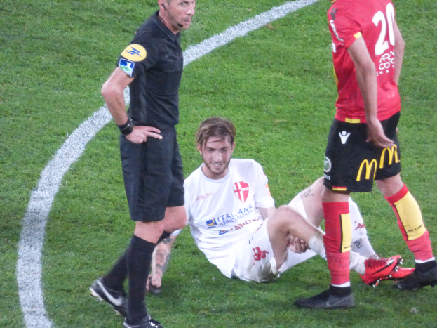 Ein Fussballspieler in einem Sportoutfit und -schuhen liegt neben einem Schiedsrichter auf dem Boden, neben ihm stehen zwei weitere Personen auf dem Rasen.