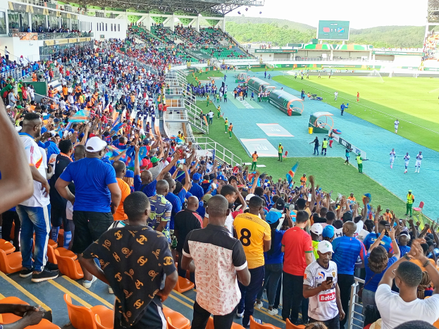 Eine große Menschenmenge versammelt sich vor einem eingezäunten Fußballfeld, wobei einige auf Stühlen sitzen und andere stehen, mit Blick auf Hügel und einen klaren blauen Himmel während der 2019 African Cup of Nations.