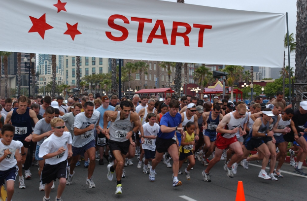 Eine Gruppe von Menschen, die bei einem Marathon laufen, mit einem Verkehrskegel im Vordergrund und einer Fahne im Hintergrund, vor Bäumen, Laternenmasten, Gebäuden und einem klaren blauen Himmel.