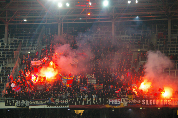 Eine große Menge Menschen in einem Stadion hält Fahnen und Banner, mit Leuchtraketen, die Rauch erzeugen, unter einer Decke mit Deckenleuchten und Metallrahmen.