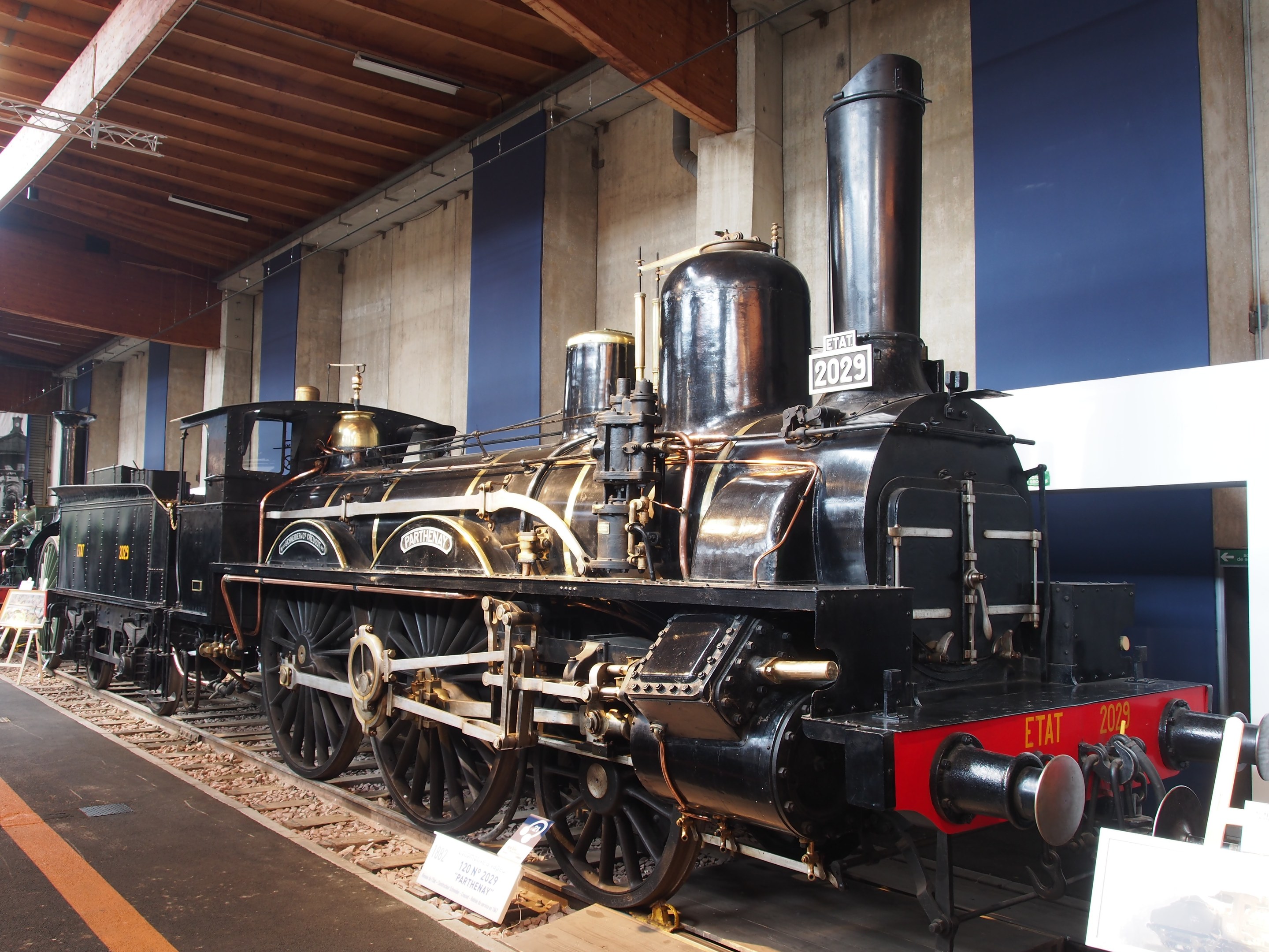 Alte Dampflokomotive in einem Museums Setting mit umgebenen Eisenbahnschienen, Informationsschildern und anderen Ausstellungen, vor einer Wand mit Deckenbeleuchtung.