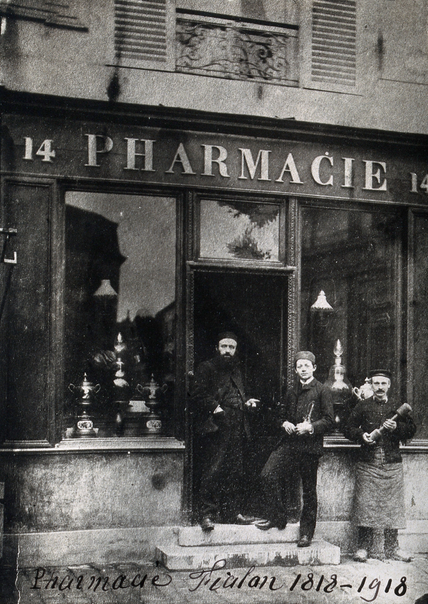 Ein altes Schwarz-Weiß-Foto von drei Männern, die vor einer Apotheke mit Fenstern, einer Tür und einem Namensschild stehen, mit verschiedenen Gegenständen im Inneren des Ladens und Text am unteren Rand des Bildes.